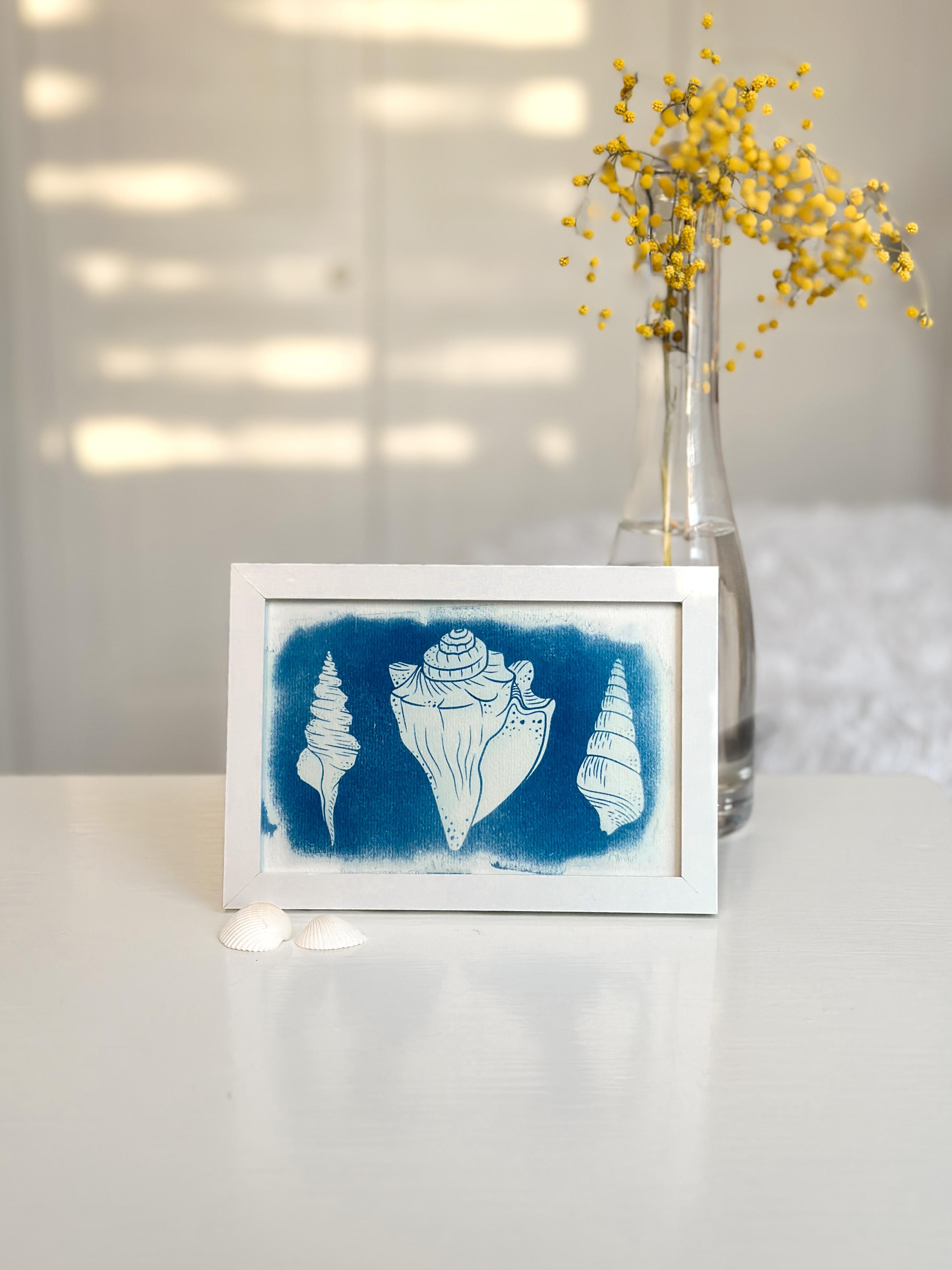 Ingelijste cyanotype print van vier schelpen, met echte schelpen en gele bloemen in vaas als decoratie
